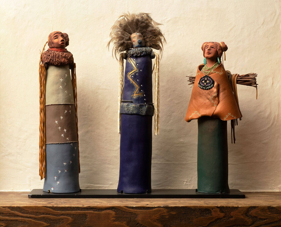 Three highly detailed cylindrical figures in traditional attire are displayed on a dark shelf against a textured wall.