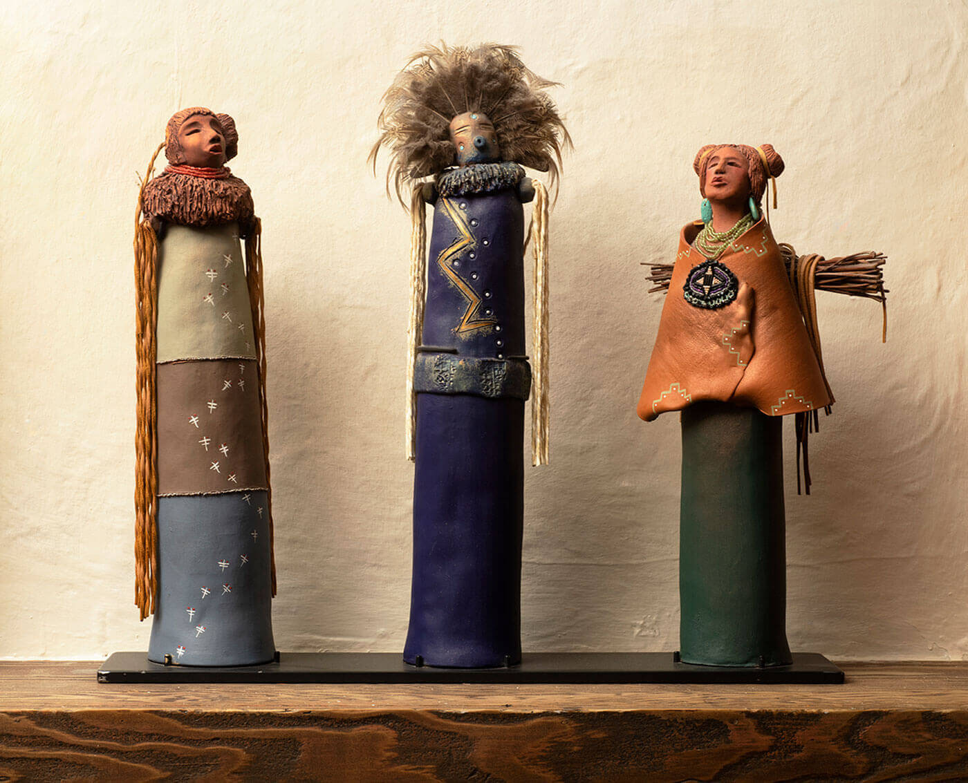 Three highly detailed cylindrical figures in traditional attire are displayed on a dark shelf against a textured wall.