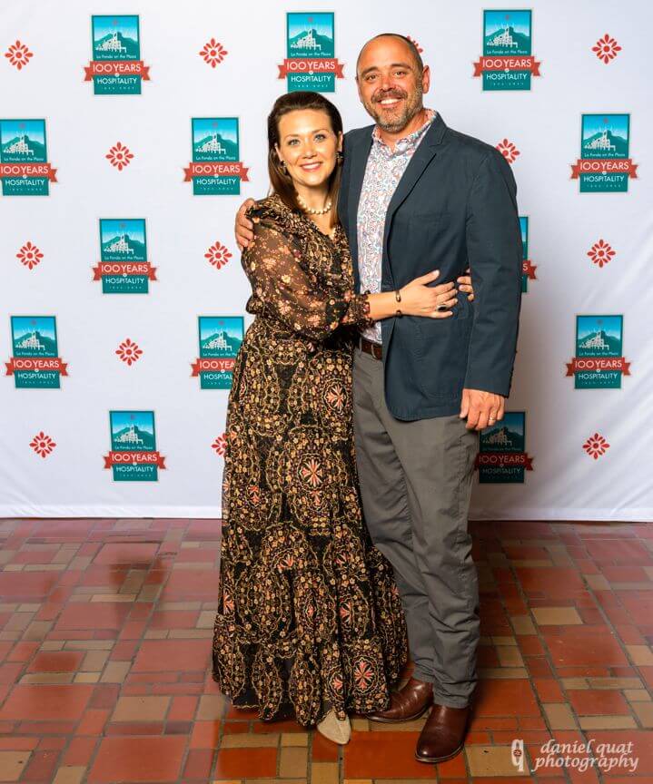 A smiling couple dressed in formal wear poses in front of the branded centennial event backdrop.