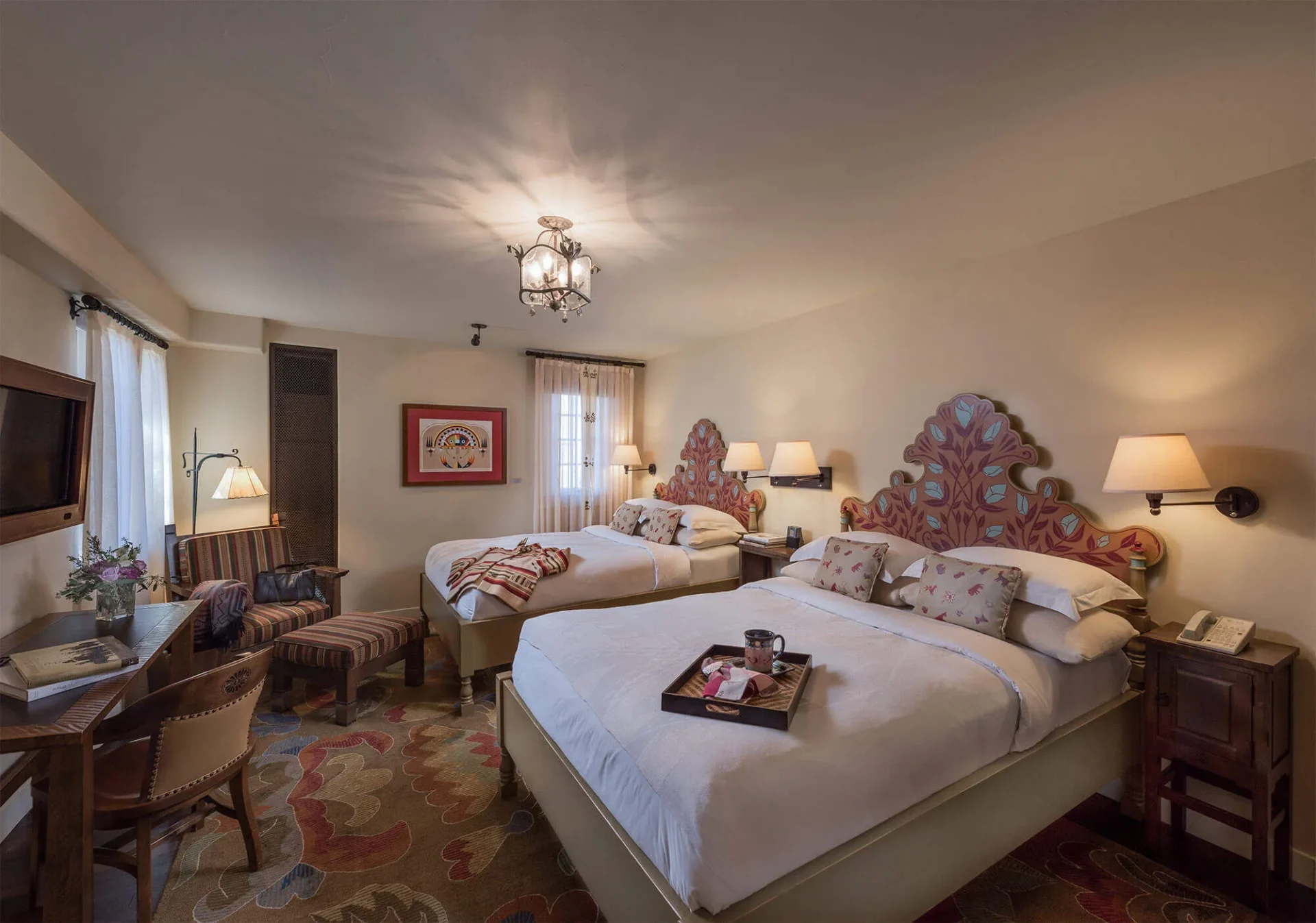 A spacious hotel room showcases two queen beds with brightly painted headboards and an intricately patterned rug.