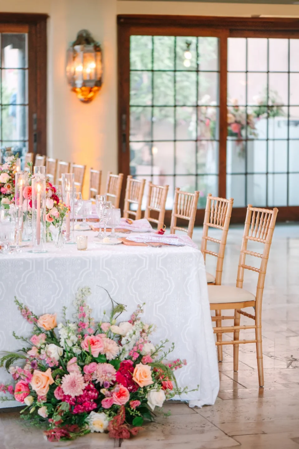 A long banquet hall dining table is beautifully set with floral arrangements, candles, and light wooden chairs.