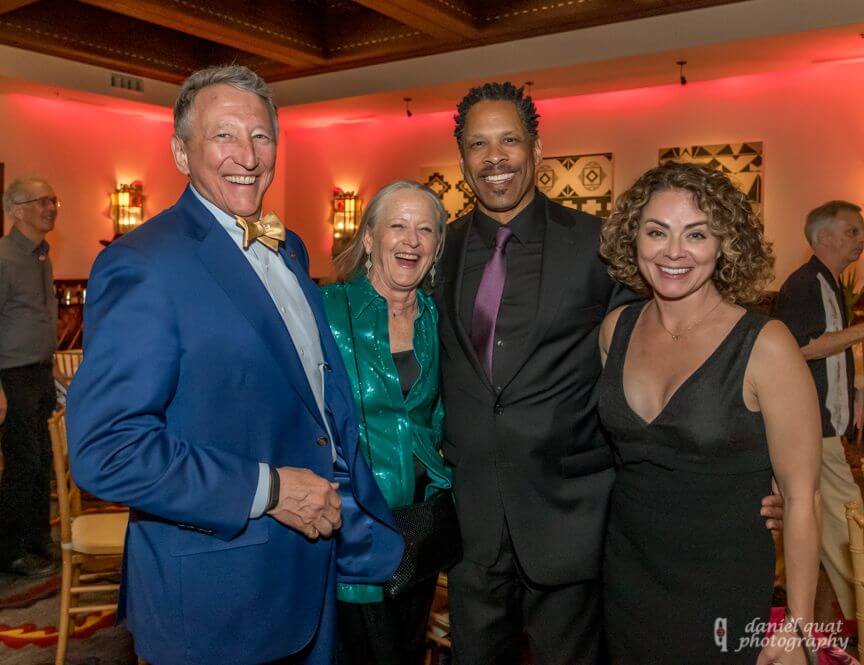 Four guests wearing formal attire smile brightly in a warm, red-lit hotel ballroom.