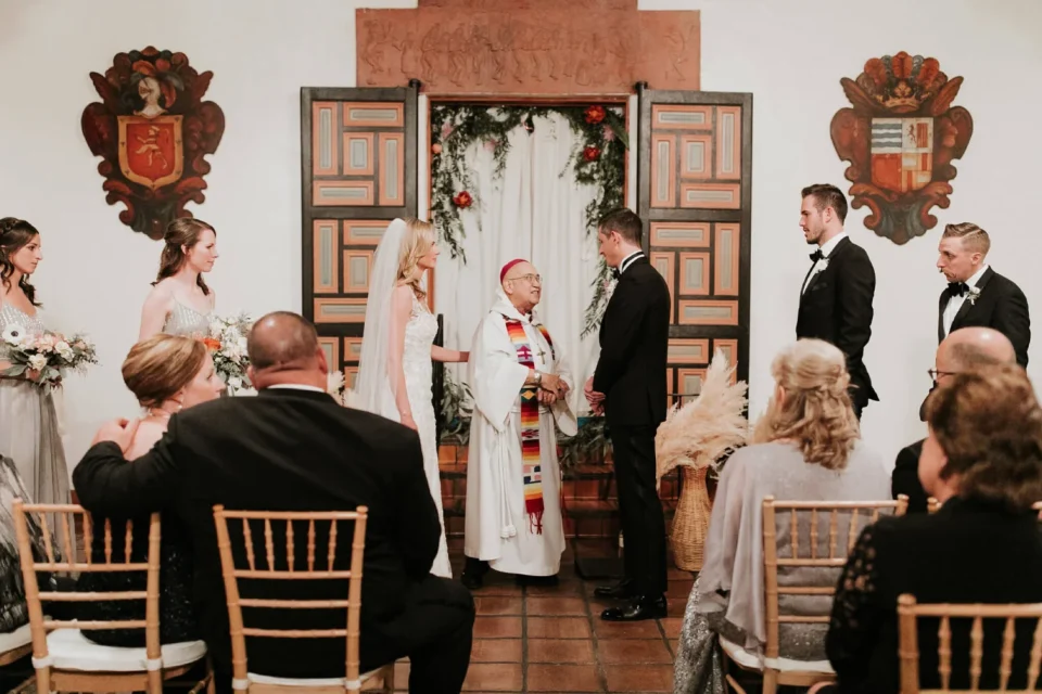 A bride and groom exchange vows before an officiant during a ceremony held in an elegant, ornate wedding venue.