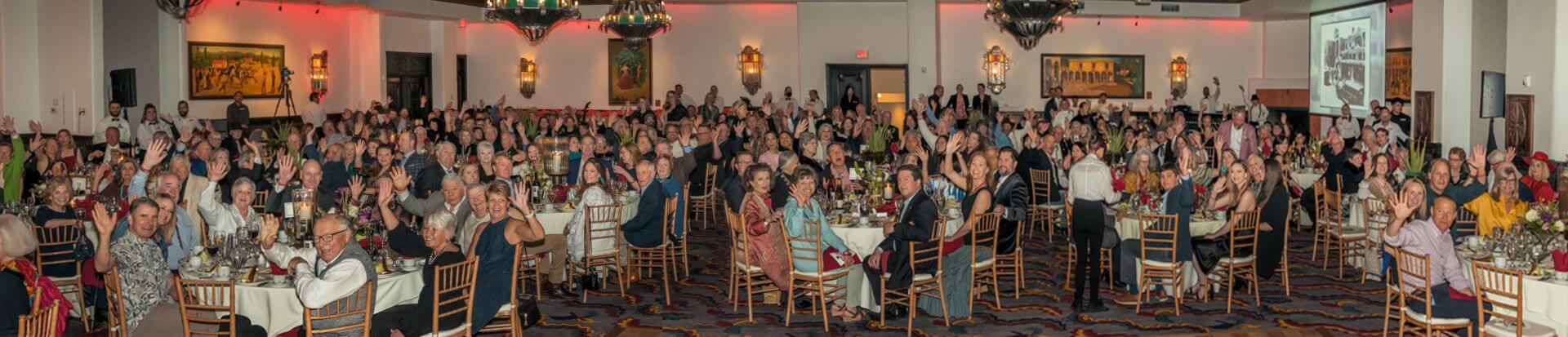 A large crowd of cheerful guests wave from their tables inside the historic ballroom and banquet hall.