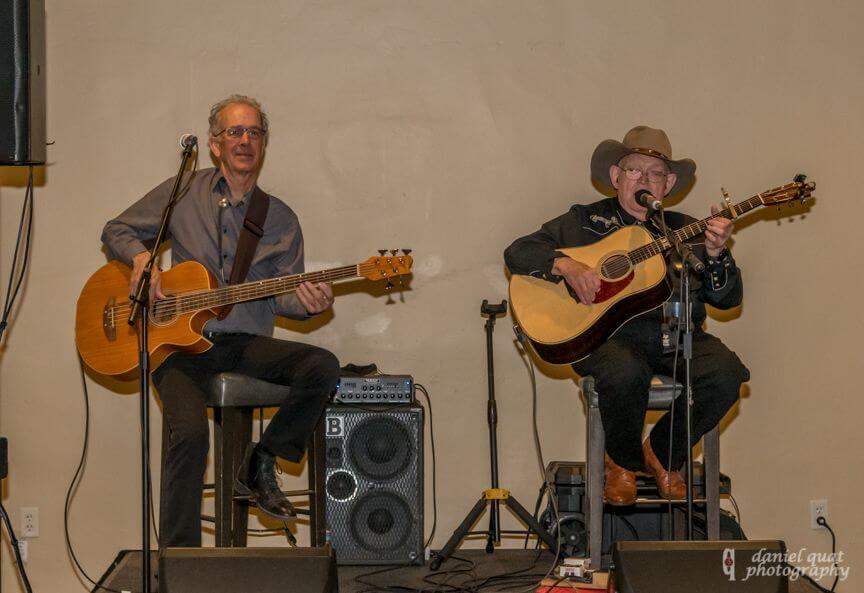 Two acoustic musicians, one wearing a cowboy hat, perform live music on stage.