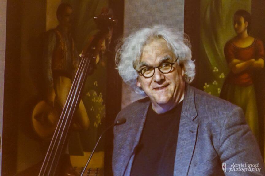 A smiling man addresses an audience next to a large upright bass, highlighting a live music event.