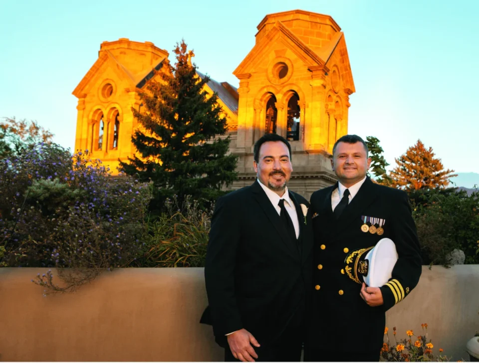 Two grooms in formal attire and military dress uniform pose outside a stunning historic building at golden hour.