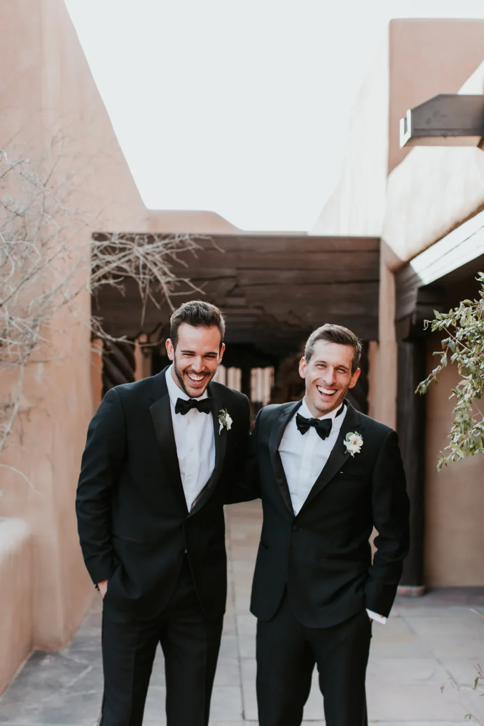 Two grooms in black tuxedos laugh happily together while standing in a beautiful Southwestern wedding venue.