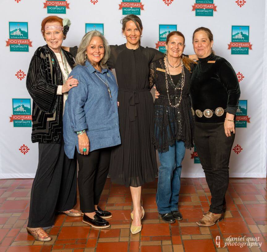 Five women dressed in event attire pose happily in front of the celebratory 100-year backdrop.