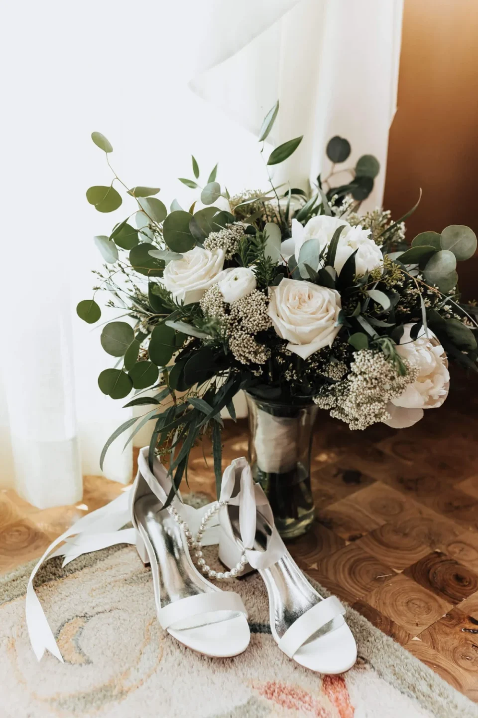 A pair of white satin high heels rests next to a large bouquet of white roses and greenery, signifying a wedding day.