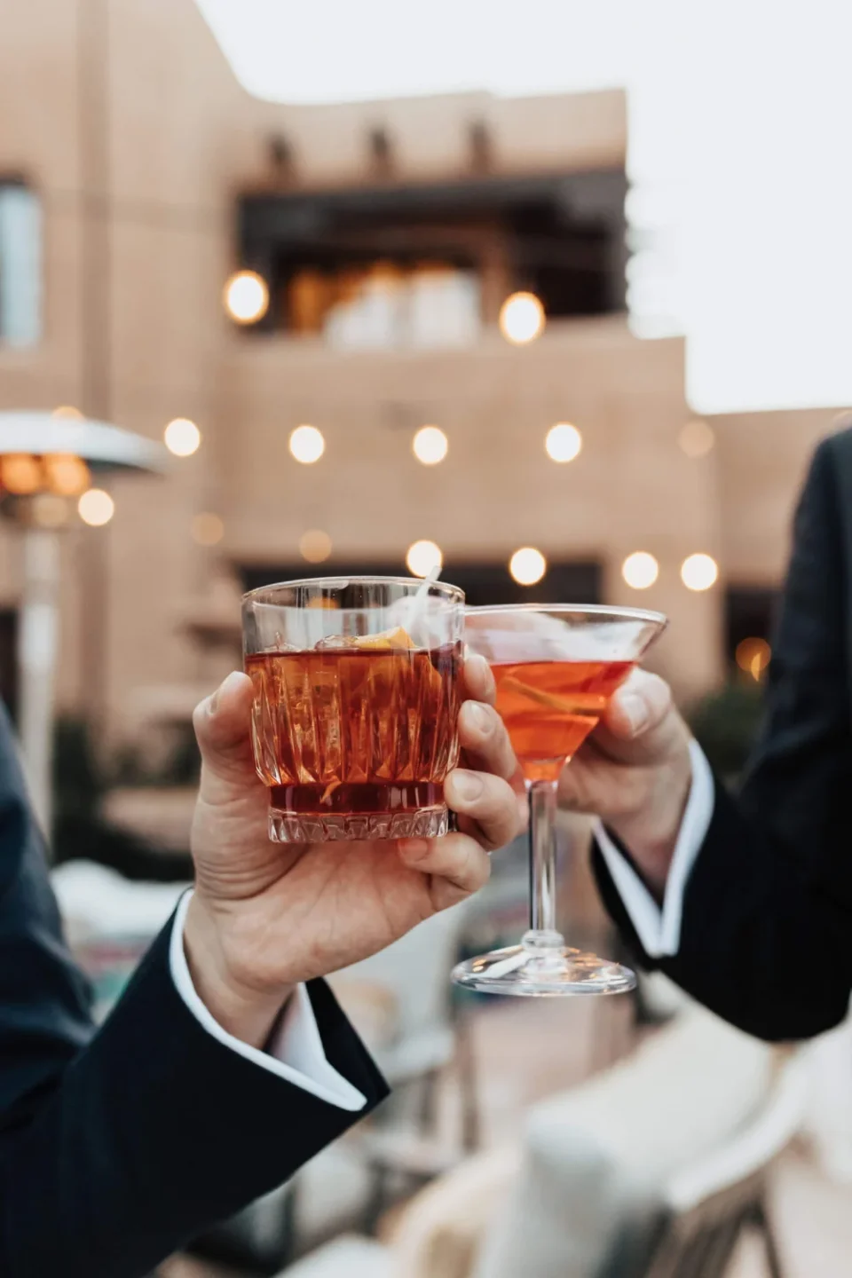 Two people toast with red cocktails on a warmly lit outdoor patio or rooftop bar.