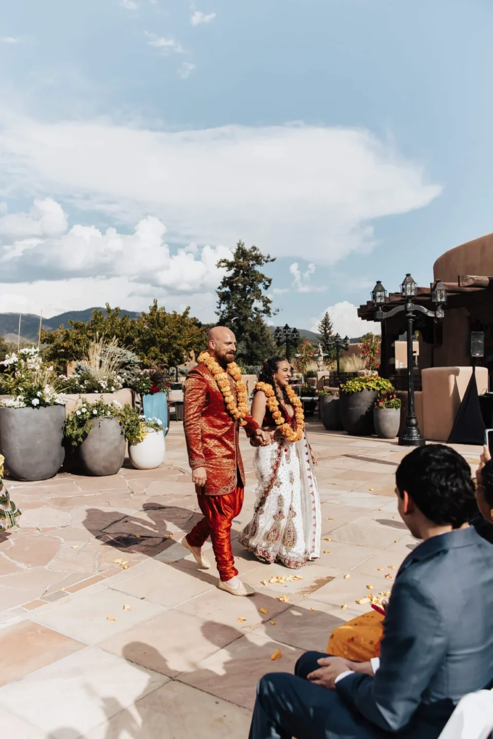 A bride and groom in traditional Indian attire walk hand-in-hand across the beautiful outdoor wedding venue patio.