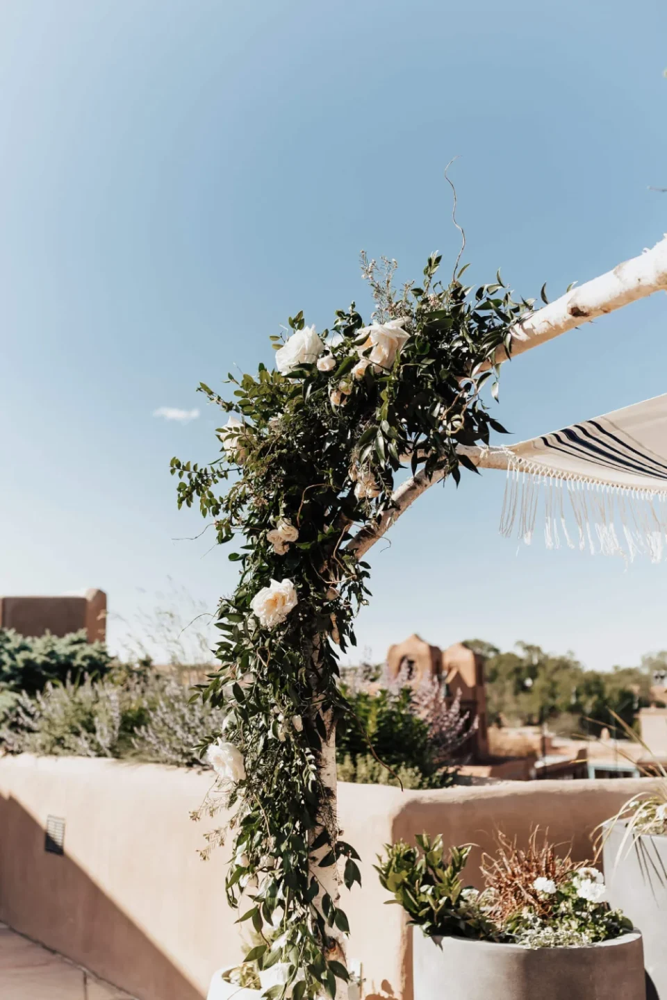 White roses and green foliage beautifully adorn a wooden arch set up for a Santa Fe wedding venue ceremony.