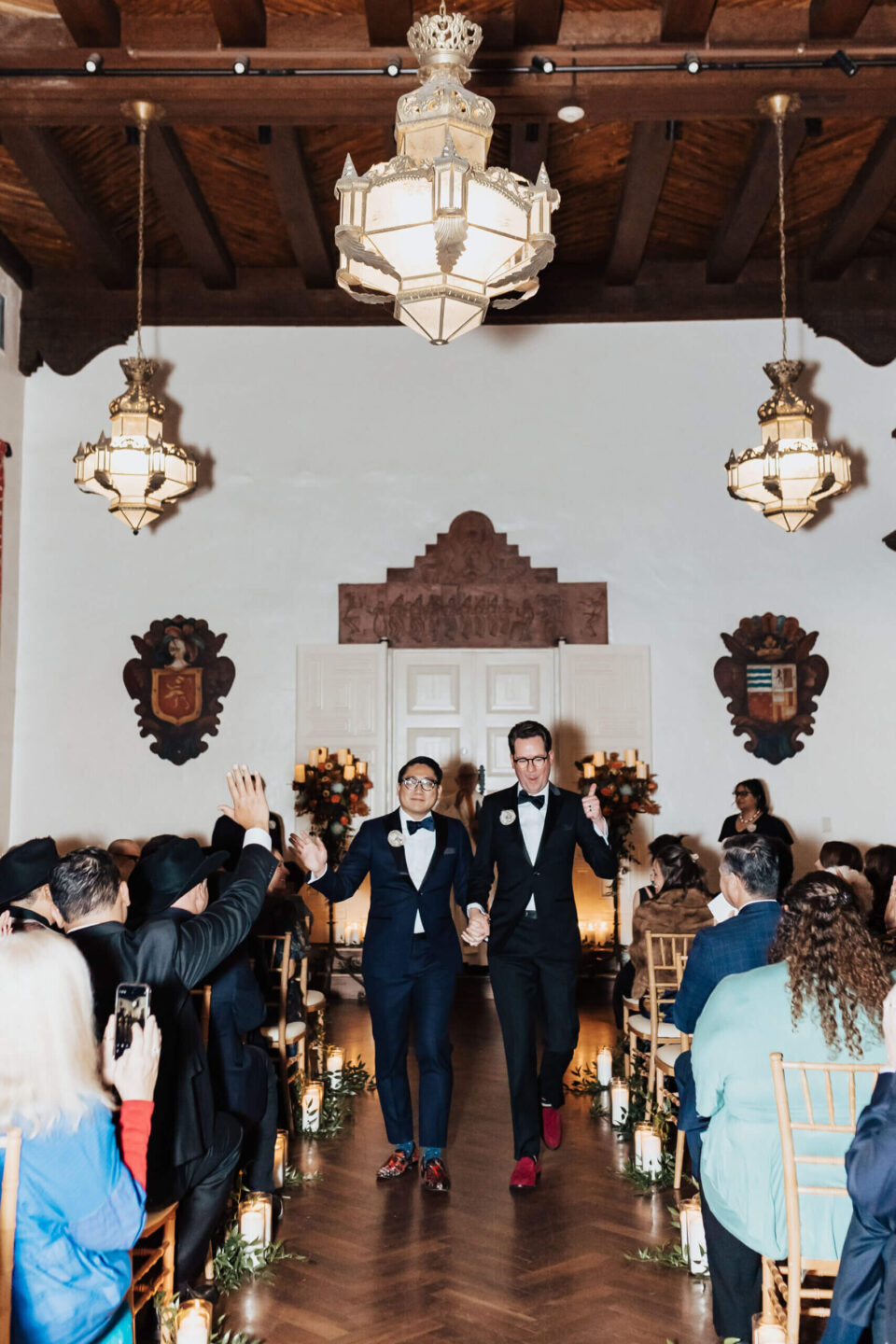 Two grooms smile and celebrate walking down the aisle of a beautifully lit ballroom ceremony space.