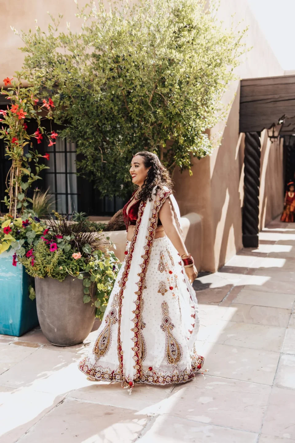 A smiling woman wearing a beautiful white and red traditional lehenga walks through the sunny outdoor event space.