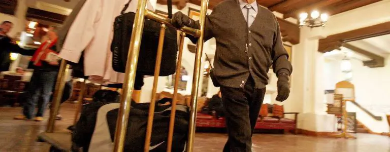 A bellman in a cardigan pushes a full brass luggage cart through the warm and traditional hotel lobby.