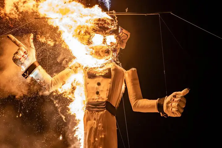 A large humanoid effigy dressed in a tuxedo is engulfed in bright orange flames against a deep black night sky.