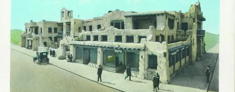 A hand-colored, vintage photo of the historic Pueblo style hotel exterior facade viewed from the street.