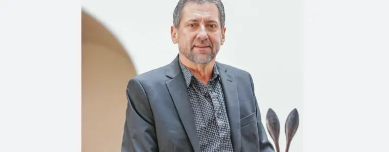 A professional man wearing a dark suit and patterned shirt stands smiling in a bright indoor setting.