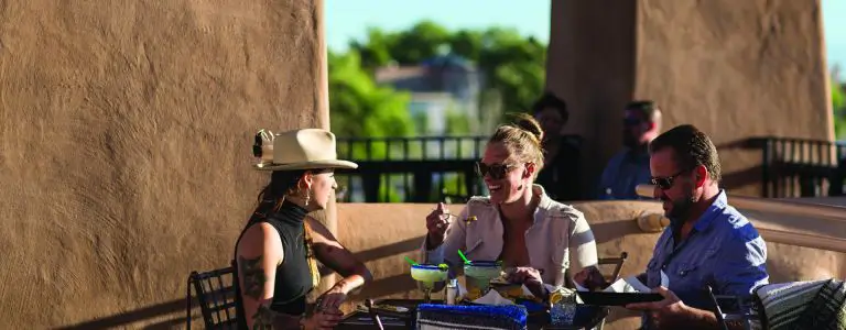 Three smiling friends enjoy drinks and appetizers together on a sunny outdoor terrace.