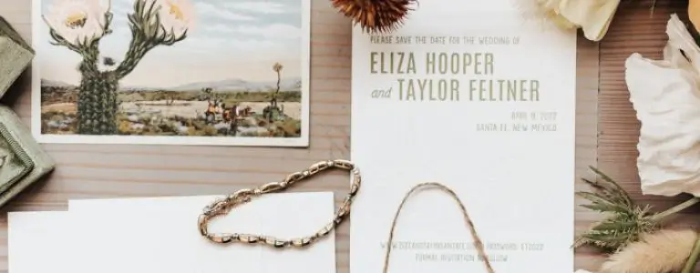 A rustic wedding invitation suite featuring a desert landscape postcard and a delicate gold chain.