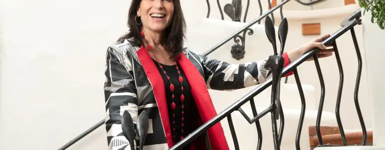 A smiling woman in a striking red and black jacket holds a wrought iron railing with a decorative rabbit design.