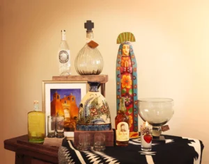 A wooden table displays colorful southwestern folk art, bottles of liquor, and framed artwork of an adobe church.