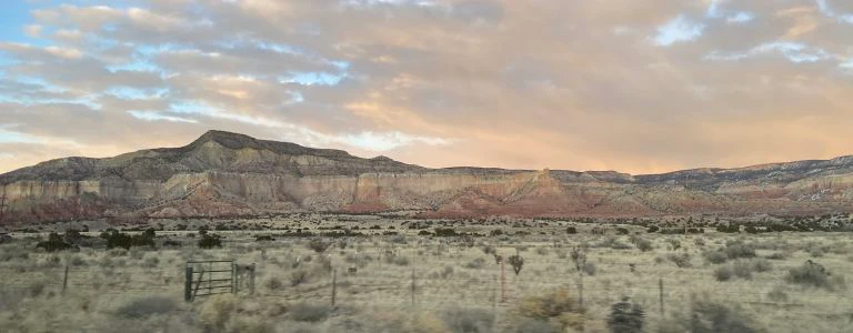 The rugged desert terrain stretches before a colorful mesa cliff face under a dramatic sunset sky.
